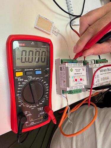 На изображении показан мультиметр с показанием 0.000 мА, подключенный к плате и датчику, на котором человек использует щупы для измерений. (Captioned by AI)