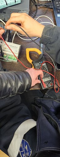 На изображении изображены руки, использующие мультиметр и токовые клещи для измерения электрических параметров на рабочем столе с различными электронными компонентами. (Captioned by AI)