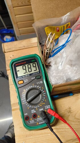 На изображении цифровой мультиметр марки Mastech, показывающий значение -9.89, рядом с коробкой, полной проводов и других предметов. (Captioned by AI)