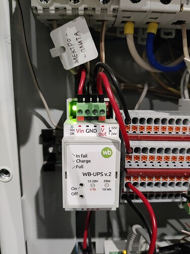 На изображении показан источник бесперебойного питания WB-UPS v.2, подключенный к электрической панели с видимыми проводами и индикаторами статуса. (Captioned by AI)